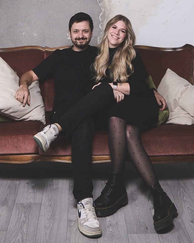 Jacky & Fabi sitzen auf einem braunen Ledersofa vor einer rauen Betonwand